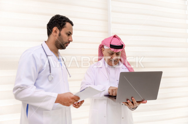 طبيبان عربيان سعوديان خليجيان يتناقشان مع بعضهم ، يستخدمان الحاسوب المحمول في العمل، العمل الجماعي للأطباء الاخصائيين في المستشفى ، مفهوم الطب والرعاية الصحية