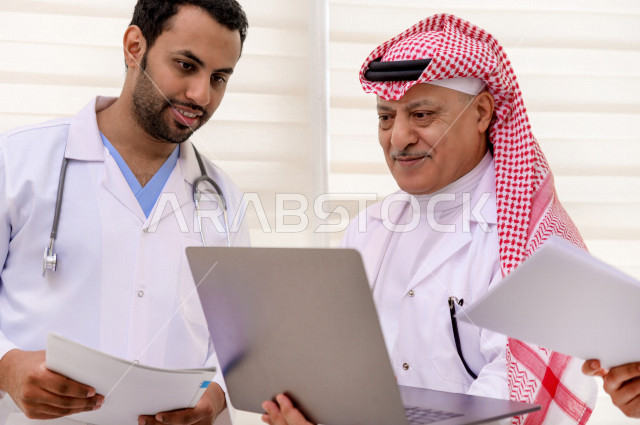 طبيبان عربيان سعوديان خليجيان يتناقشان مع بعضهم ، يستخدمان الحاسوب المحمول في العمل، العمل الجماعي للأطباء الاخصائيين في المستشفى ، مفهوم الطب والرعاية الصحية