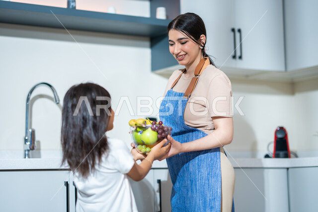 ايماءات تدل على الفرح والسعادة, صورة من الخلف لفتاة عربية خليجية سعودية مع والدتها في المطبخ, تقديم وعاء من الفواكه المتنوعة الطازجة, الفاكهة اللذيذة الشهية, مفهوم النظام الغذائي, غذاء صحي ومفيد