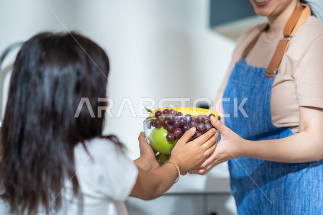 الفاكهة اللذيذة الشهية, صورة مقربة  من الخلف لفتاة عربية خليجية سعودية مع والدتها في المطبخ, تقديم وعاء من الفواكه المتنوعة الطازجة, مفهوم النظام الغذائي, غذاء صحي ومفيد