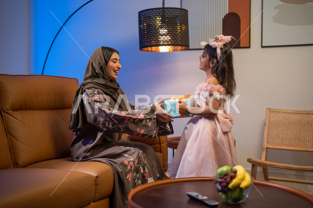 مفاجآت سعيدة في الاعياد والمناسبات , إيماءات تدل على السعادة بالهدايا و التهنئات, صورة من الخلف لفتاة عربية خليجية سعودية تقوم بتقديم علبة هدية مغلفة لوالدتها, إيماءات وجه تدل على الفرح والسعادة بالهدايا و التهنئات