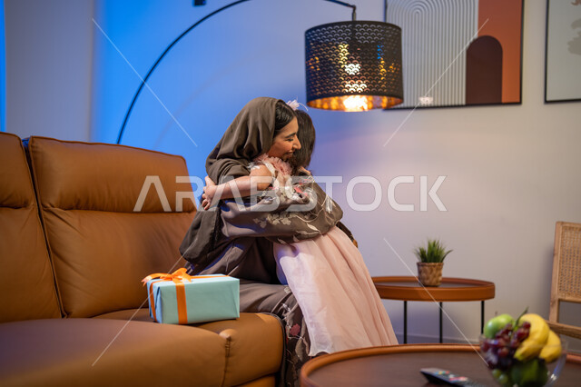  أجواء العيد في المجتمع العربي السعودي, تقديم الهدايا المغلفة ,أم عربية خليجية سعودية ترتدي العباءة الخليجية في غرفة المعيشة تقوم بضم وحضن ابنتها, ايماءات تدل على السعادة بالمفاجآت, أعياد ومناسبات اسلامية