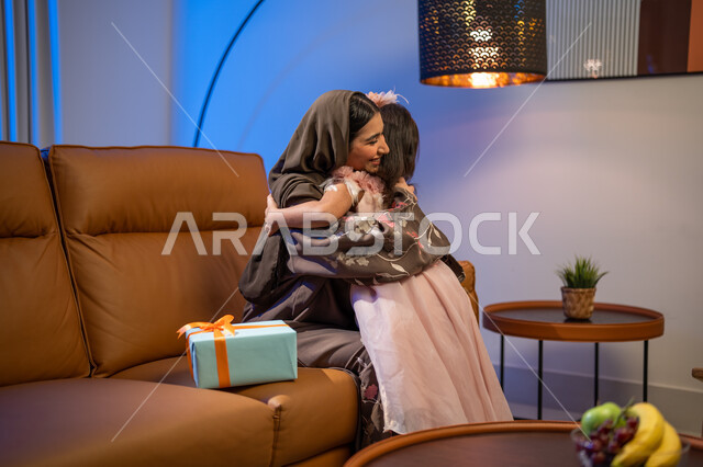  أجواء العيد في المجتمع العربي السعودي, تقديم الهدايا المغلفة ,أم عربية خليجية سعودية ترتدي العباءة الخليجية في غرفة المعيشة تقوم بضم وحضن ابنتها, ايماءات تدل على السعادة بالمفاجآت, أعياد ومناسبات اسلامية