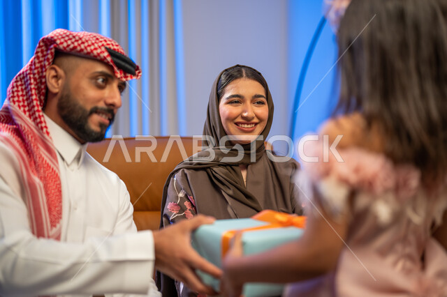 أجواء العيد في المجتمع العربي السعودي, صورة من الخلف لفتاة عربية خليجية سعودية تقوم بتقديم علبة هدية مغلفة لوالدها في غرفة المعيشة, إيماءات وجه تدل على الفرح والسعادة بالهدايا و التهنئات, أعياد ومناسبات اسلامية