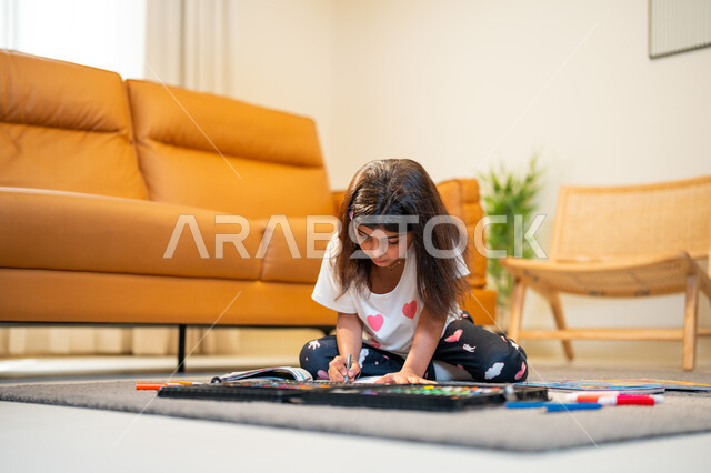 ادوات ومعدات التلوين، التعلم والابداع وتنمية المهارات، طفلة عربية خليجية سعودية تجلس في غرفة المعيشة وتبدع بتلوين الصور، قضاء اوقات ممتعة، لعبة تلوين الرسومات، ايماءات وجه تدل على السعادة، انشطة ترفيهية فنية ابداعية
