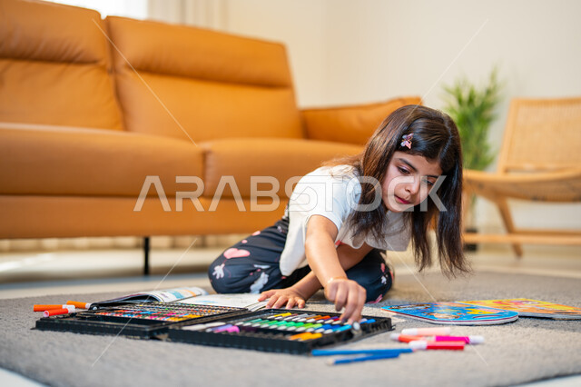الابداع بتلوين الصور من خلال ادوات ومعدات التلوين، طفلة عربية خليجية سعودية تجلس في غرفة المعيشة، التعلم والابداع وتنمية المهارات، قضاء اوقات ممتعة، لعبة تلوين الرسومات، ايماءات وجه تدل على السعادة، انشطة ترفيهية فنية ابداعية