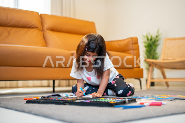 الابداع بتلوين الصور من خلال ادوات ومعدات التلوين، طفلة عربية خليجية سعودية تجلس في غرفة المعيشة، التعلم والابداع وتنمية المهارات، قضاء اوقات ممتعة، لعبة تلوين الرسومات، ايماءات وجه تدل على السعادة، انشطة ترفيهية فنية ابداعية