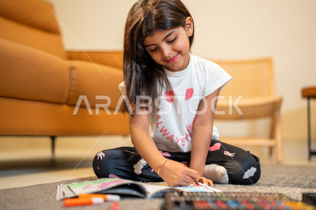 التعلم والابداع وتنمية المهارات، طفلة عربية خليجية سعودية تجلس في غرفة المعيشة وتبدع بتلوين الصور، ادوات ومعدات التلوين، قضاء اوقات ممتعة، لعبة تلوين الرسومات، ايماءات وجه تدل على السعادة، انشطة ترفيهية فنية ابداعية