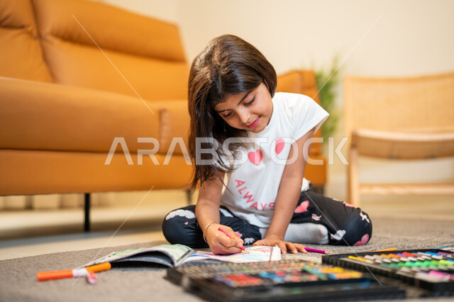 انشطة ترفيهية فنية ابداعية، طفلة عربية خليجية سعودية تجلس في غرفة المعيشة وتبدع بتلوين الصور، ادوات ومعدات التلوين، التعلم والابداع وتنمية المهارات، قضاء اوقات ممتعة، لعبة تلوين الرسومات، ايماءات وجه تدل على السعادة
