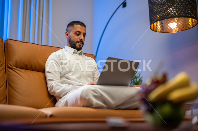 عقد إجتماع عمل عبر الإنترنت، إستخدام جهاز الحاسوب المحمول، رجل عربي خليجي سعودي يجلس في غرفة المعيشة ،التسويق لموقع إلكتروني، إجراء مكالمة فيديو، مشاهدة الأفلام والمسلسلات