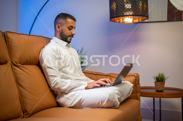 Marketing a website, using a laptop, a Saudi Arabian Gulf man sitting in the living room, holding a business meeting via the Internet, making a video call, watching movies and series