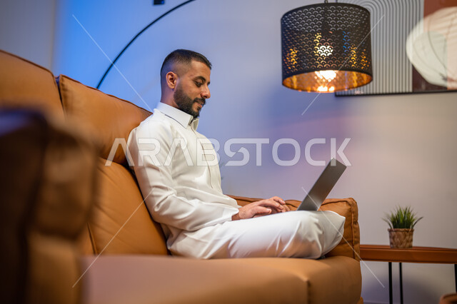 إستخدام جهاز الحاسوب المحمول، رجل عربي خليجي سعودي يجلس في غرفة المعيشة ،عقد إجتماع عمل عبر الإنترنت، التسويق لموقع إلكتروني، إجراء مكالمة فيديو، مشاهدة الأفلام والمسلسلات