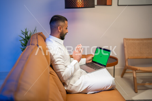 ايماءات تدل على التحية والسلام, إجراء مكالمة فيديو، استخدام جهاز الحاسوب المحمول بخلفية خضراء كروما، رجل عربي خليجي سعودي يجلس في غرفة المعيشة ، التسويق لموقع إلكتروني، مشاهدة الأفلام والمسلسلات