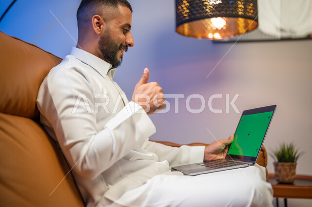 Gestures indicating the quality mark, using a laptop with a green chroma background, a Saudi Arabian Gulf man sitting in the living room, making a video call, marketing a website, watching movies and series