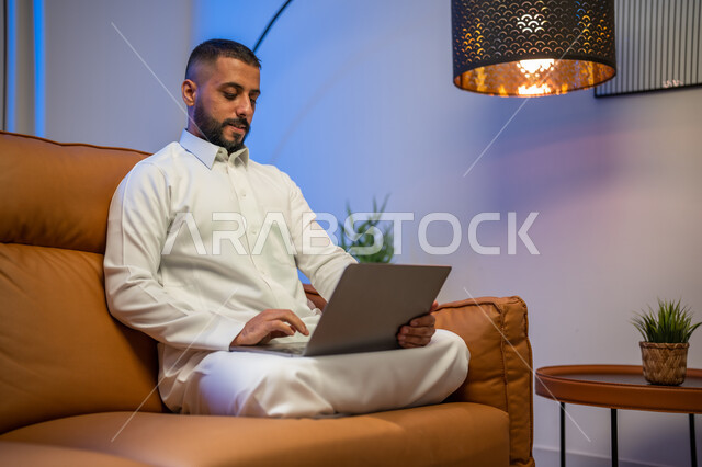 إستخدام جهاز الحاسوب المحمول، رجل عربي خليجي سعودي يجلس في غرفة المعيشة ،عقد إجتماع عمل عبر الإنترنت، التسويق لموقع إلكتروني، إجراء مكالمة فيديو، مشاهدة الأفلام والمسلسلات