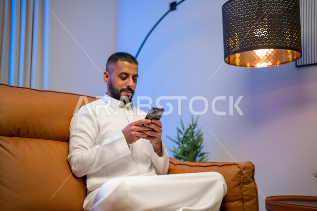 The use of social media and modern technologies, a Saudi Arabian Gulf man sitting in the living room using a mobile phone, surfing the Internet via mobile, communicating with friends