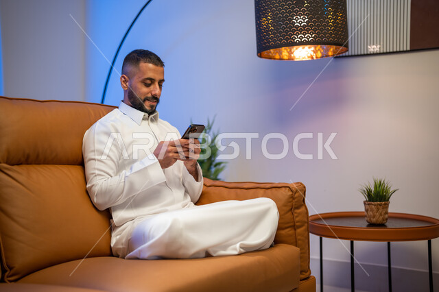 تصفح الإنترنت عبر الجوال، إستخدام وسائل التواصل الإجتماعي و التقنيات الحديثة, رجل عربي خليجي سعودي  يجلس في غرفة المعيشة يستخدم الهاتف المحمول, التواصل مع الأصدقاء