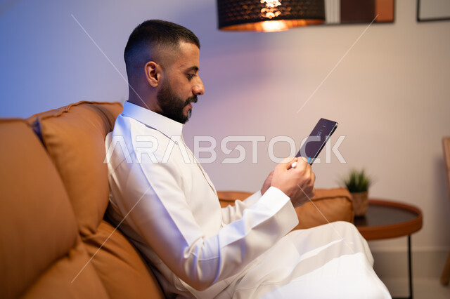 برامج التواصل الاجتماعي, رجل عربي خليجي سعودي يجلس في غرفة المعيشة يستخدم التابليت المحمول، التسويق لموقع إلكتروني ، استخدام قلم التابليت لتدوين الملاحظات، التقنيات الحديثة المتطورة