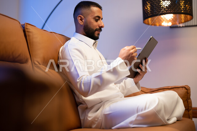 التقنيات الحديثة المتطورة، رجل عربي خليجي سعودي يجلس في غرفة المعيشة يستخدم التابليت المحمول، التسويق لموقع إلكتروني ، استخدام قلم التابليت لتدوين الملاحظات، برامج التواصل الاجتماعي