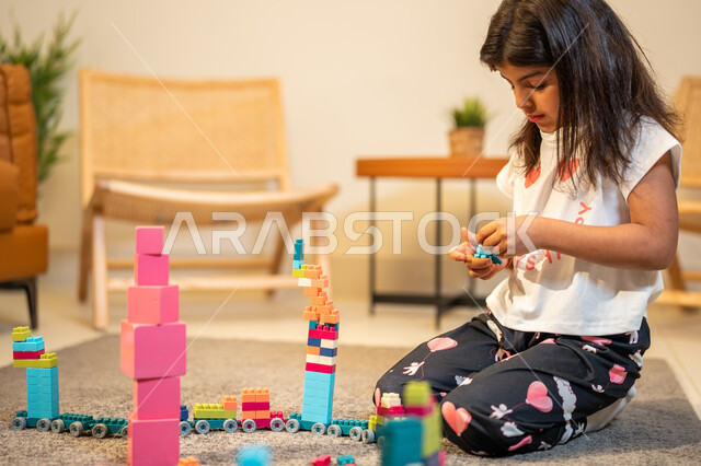 انشطة ترفيهية ابداعية، طفلة عربية خليجية سعودية تستمتع بتركيب المكعبات، الجلوس في غرفة المعيشة، ايماءات وجه تدل على السعادة، الابداع وتنمية المهارات، قضاء اوقات ممتعة