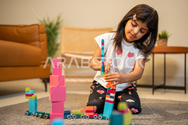انشطة ترفيهية ابداعية، طفلة عربية خليجية سعودية تستمتع بتركيب المكعبات، الجلوس في غرفة المعيشة، ايماءات وجه تدل على السعادة، الابداع وتنمية المهارات، قضاء اوقات ممتعة
