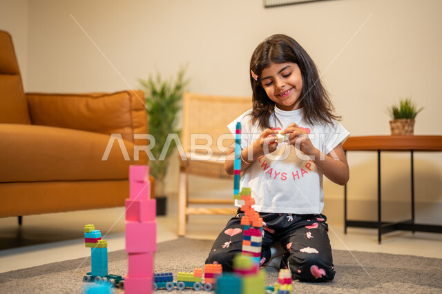 الابداع وتنمية المهارات، طفلة عربية خليجية سعودية تستمتع بتركيب المكعبات، الجلوس في غرفة المعيشة، ايماءات وجه تدل على السعادة، انشطة ترفيهية ابداعية، قضاء اوقات ممتعة