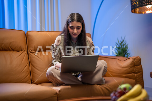 العمل عن بعد, إجراء مكالمة فيديو , شابة عربية خلجية سعودية تجلس في غرفة المعيشة, استخدام الحاسوب المحمول, الدردشة و التواصل الاجتماعي, تصفح الانترنت واستخدام تقنيات حديثة, التسويق لموقع الكتروني