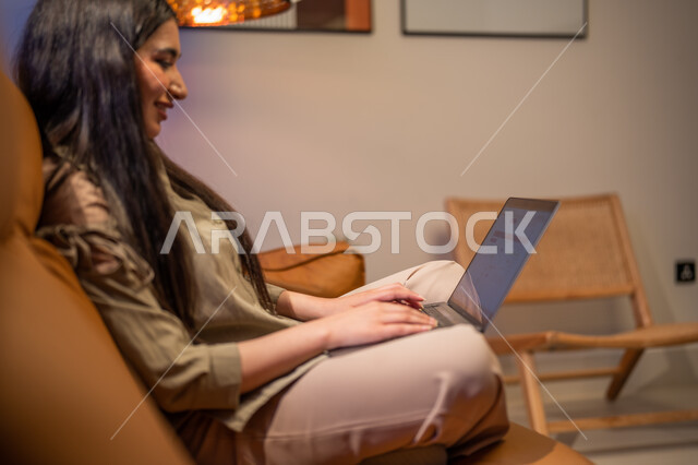 ايماءات تدل على السعادة, إجراء مكالمة فيديو , العمل عن بعد, شابة عربية خلجية سعودية تجلس في غرفة المعيشة, استخدام الحاسوب المحمول, الدردشة و التواصل الاجتماعي, تصفح الانترنت واستخدام تقنيات حديثة, التسويق لموقع الكتروني