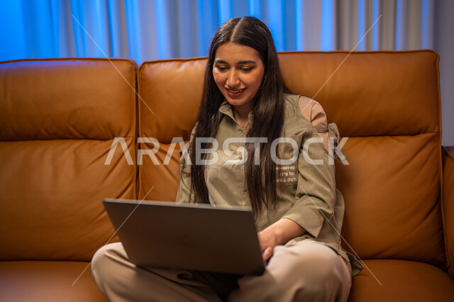 ايماءات تدل على السعادة, العمل عن بعد, إجراء مكالمة فيديو ,  شابة عربية خلجية سعودية تجلس في غرفة المعيشة, استخدام الحاسوب المحمول, الدردشة و التواصل الاجتماعي, تصفح الانترنت واستخدام تقنيات حديثة, التسويق لموقع الكتروني
