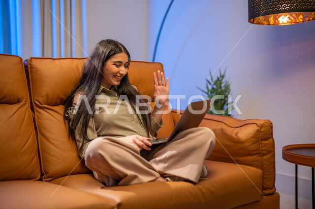 إجراء مكالمة فيديو , ايماءات تدل على التحية والسلام, شابة عربية خلجية سعودية تجلس في غرفة المعيشة, استخدام الحاسوب المحمول, الدردشة و التواصل الاجتماعي, تصفح الانترنت واستخدام تقنيات حديثة, بث مباشر