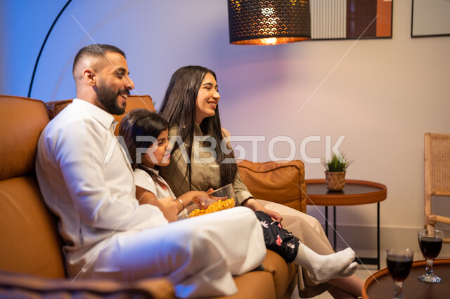 Watching movies and series, gestures indicating happiness and joy, enjoying eating popcorn, a family atmosphere full of love, a Saudi Arabian Gulf family in the living room, spending quality time with children