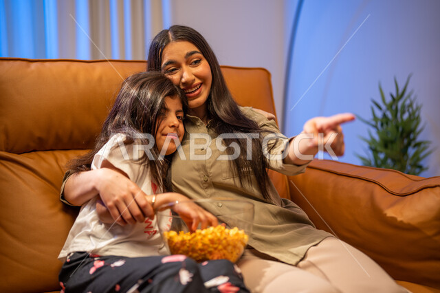 الاشارة الى شيئ ما, الإهتمام والاعتناء بالأبناء, أجواء عائليه مليئة بالحب, أم عربيه سعوديه خليجيه تقوم  بحضن وضم ابنتها في غرفة المعيشة, ايماءات تدل على السعادة و الفرح , قضاء أوقات ممتعه مع الأبناء , الاستمتاع بتناول الفوشار