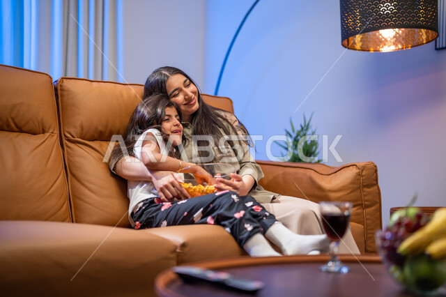 الإهتمام والاعتناء بالأبناء, أجواء عائليه مليئة بالحب, أم عربيه سعوديه خليجيه تقوم  بحضن وضم ابنتها في غرفة المعيشة, ايماءات تدل على السعادة و الفرح , قضاء أوقات ممتعه مع الأبناء , الاستمتاع بتناول الفوشار