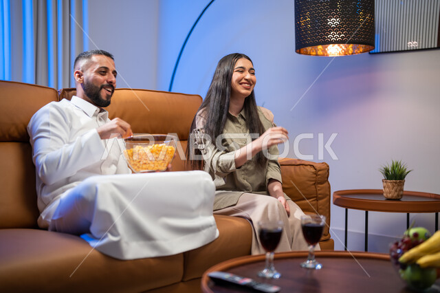 تناول المقرمشات والمسليات، زوجان خليجيان سعوديان عربيان يقضيان اوقات ممتعة، الجلوس في غرفة المعيشة، مشاهدة المسلسلات والبرامج التلفزيونية، ايماءات وجه تدل على السعادة، الاستمتاع باوقات الفراغ، اجواء عائلية سعيدة