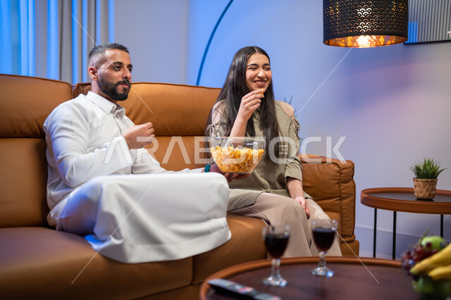 ايماءات تدل على الفرح و السرور, المحادثة والدردشات بين أفراد العائلة, الاستمتاع بأوقات الفراغ وتناول الفوشار, زوجان عربيان خليجيان سعوديان في غرفة المعيشة, أجواء عائلية مليئة بالحب, مشاهدة الأفلام والمسلسلات