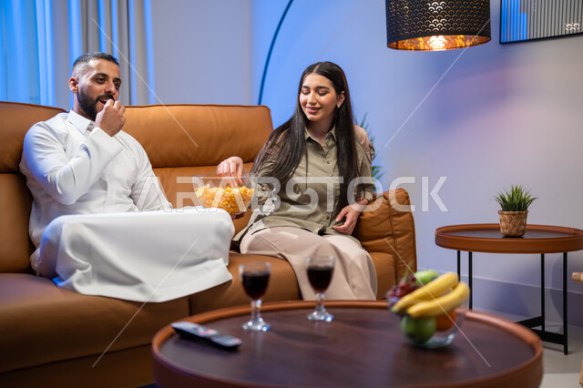 ايماءات تدل على الفرح و السرور, المحادثة والدردشات بين أفراد العائلة, الاستمتاع بأوقات الفراغ وتناول الفوشار, زوجان عربيان خليجيان سعوديان في غرفة المعيشة, أجواء عائلية مليئة بالحب, مشاهدة الأفلام والمسلسلات