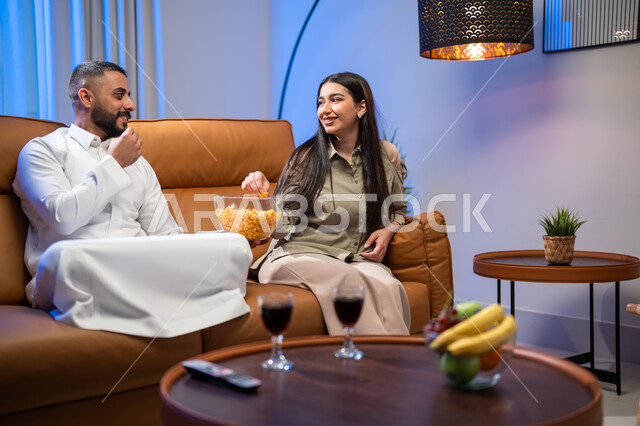 Chatting and conversations between family members, enjoying leisure time and eating popcorn, a Saudi Arabian Gulf couple in the living room, gestures indicating happiness, a family atmosphere full of love, watching movies and series