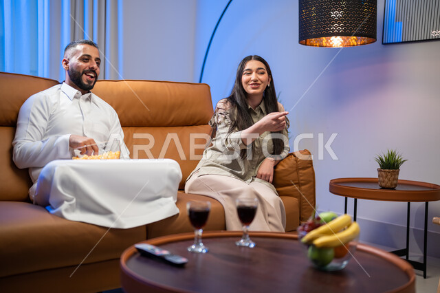 الدردشة والمحادثات بين أفراد العائلة, الاستمتاع بأوقات الفراغ وتناول الفوشار, زوجان عربيان خليجيان سعوديان في غرفة المعيشة, ايماءات تدل على السعادة, أجواء عائلية مليئة بالحب, مشاهدة الأفلام والمسلسلات