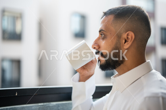رجل عربي خليجي سعودي يقف امام النافذة ويسمك كوب، الاستمتاع بالمشروبات الساخنة، فنجان زجاجي ابيض، قضاء الوقت باحتساء مشروب ساخن