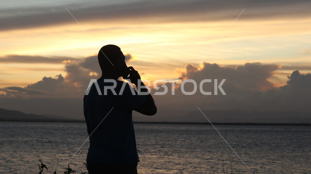 استخدام أجهزة تقنية حديثة, الاسترخاء والتأمل , صورة ظلية من الخلف لشاب يقف أمام بحيرة مائية ويستخدم الهاتف المحمول , السماء والمناظر الطبيعية , منظر غروب الشمس ، خلفية الطبيعة , سوليليت .