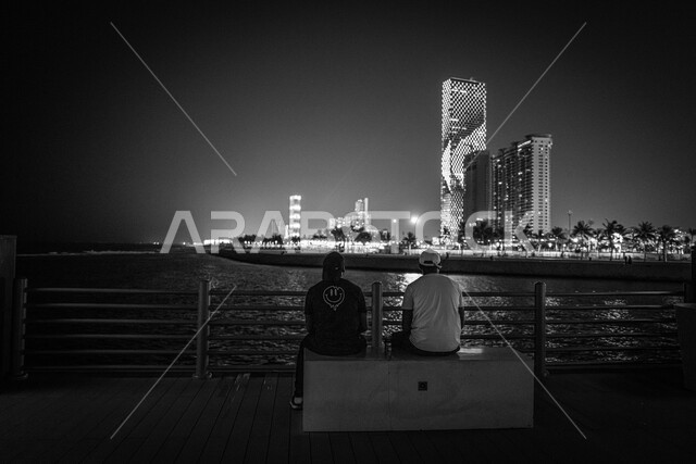 Enjoying with friends, the waterfront of Jeddah Corniche in the Kingdom of Saudi Arabia at night, a black and white picture of two young men contemplating the beauty and tranquility of the sea, the towers and skyscrapers, the famous tourist attractions of