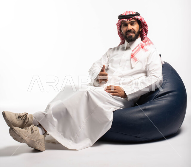 Looking at the camera, a portrait of a Saudi Arabian Gulf man wearing traditional dress sitting on a comfortable chair, hand gestures and movements indicating admiration and acceptance, white background