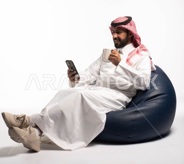 قضاء وقت ممتع, بورتريه لرجل عربي خليجي سعودي يجلس على كرسي مريح يحمل بيده فنجان القهوة ويستخدم الهاتف المحمول ، استخدام البرامج والتطبيقات الالكترونية، التسوق الالكتروني عبر الانترنت، تصفح المواقع الالكترونية والتواصل الاجتماعي، خلفية بيضاء