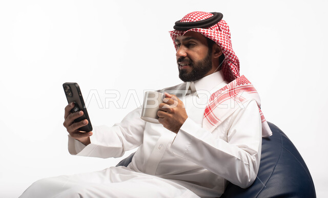 قضاء وقت ممتع, بورتريه لرجل عربي خليجي سعودي يجلس على كرسي مريح يحمل بيده فنجان القهوة ويستخدم الهاتف المحمول ، استخدام البرامج والتطبيقات الالكترونية، التسوق الالكتروني عبر الانترنت، تصفح المواقع الالكترونية والتواصل الاجتماعي، خلفية بيضاء