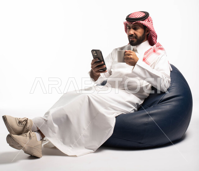 قضاء وقت ممتع, بورتريه لرجل عربي خليجي سعودي يجلس على كرسي مريح يحمل بيده فنجان القهوة ويستخدم الهاتف المحمول ، استخدام البرامج والتطبيقات الالكترونية، التسوق الالكتروني عبر الانترنت، تصفح المواقع الالكترونية والتواصل الاجتماعي، خلفية بيضاء