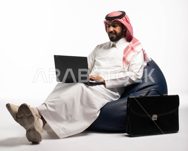 دردشة ومحادثات و تواصل اجتماعي ، التسوق الالكتروني وطلبات الاونلاين ، بورتريه لرجل عربي سعودي خليجي يرتدي الزي السعودي يجلس على كرسي مريح يستخدم الحاسوب المحمول، إنجاز مهام العمل عن بعد ، خلفية بيضاء