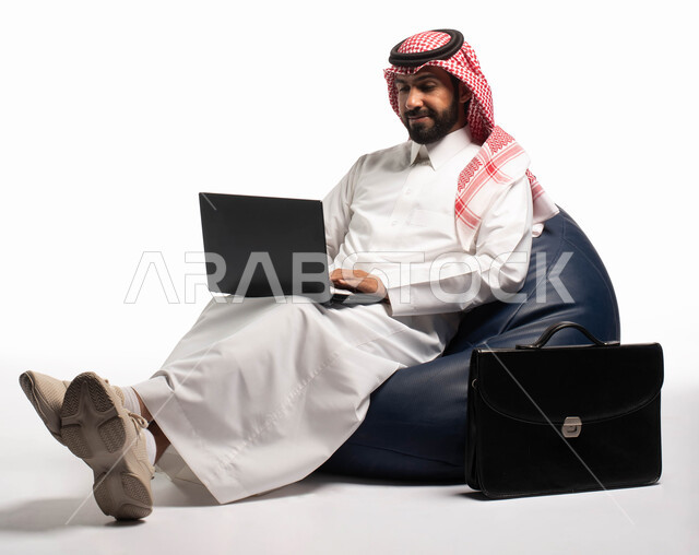 إنجاز مهام العمل عن بعد، دردشة ومحادثات و تواصل اجتماعي ، التسوق الالكتروني وطلبات الاونلاين ، بورتريه لرجل عربي سعودي خليجي يرتدي الزي السعودي يجلس على كرسي مريح يستخدم الحاسوب المحمول، خلفية بيضاء