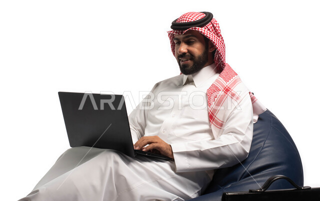 دردشة ومحادثات و تواصل اجتماعي ، إنجاز مهام العمل عن بعد، التسوق الالكتروني وطلبات الاونلاين ، بورتريه مقرب لرجل عربي سعودي خليجي يرتدي الزي السعودي يجلس على كرسي مريح يستخدم الحاسوب المحمول، خلفية بيضاء