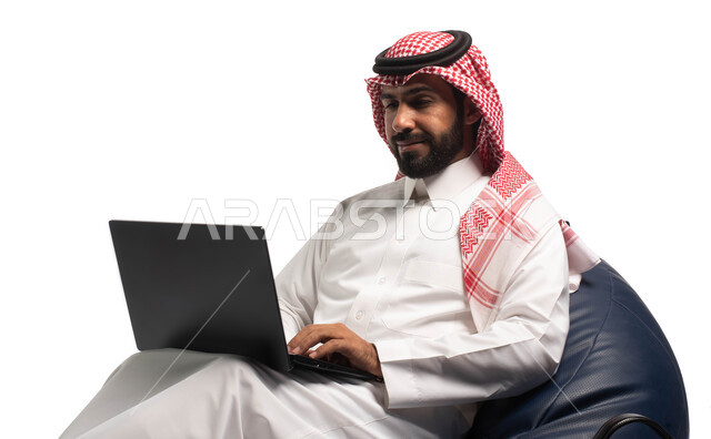 العمل عن بعد باستخدام الحاسوب المحمول ، بث مباشر واجراء مكالمة فيديو عبر جهاز الكمبيوتر ، بورتريه مقرب  لرجل عربي سعودي خليجي يرتدي الزي السعودي يجلس على كرسي مريح ، استخدام الحاسوب المحمول ، خلفية بيضاء