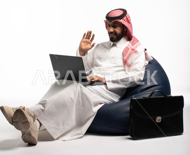 Completion of remote work tasks, chatting, conversations and social communication, online shopping and online requests, a portrait of a Saudi Arabian Gulf man wearing Saudi dress sitting on a comfortable chair using a laptop, white background